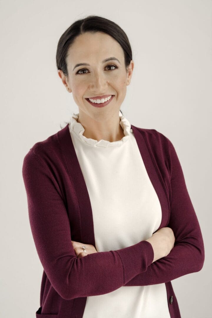 A woman in white shirt and maroon sweater standing with arms crossed.